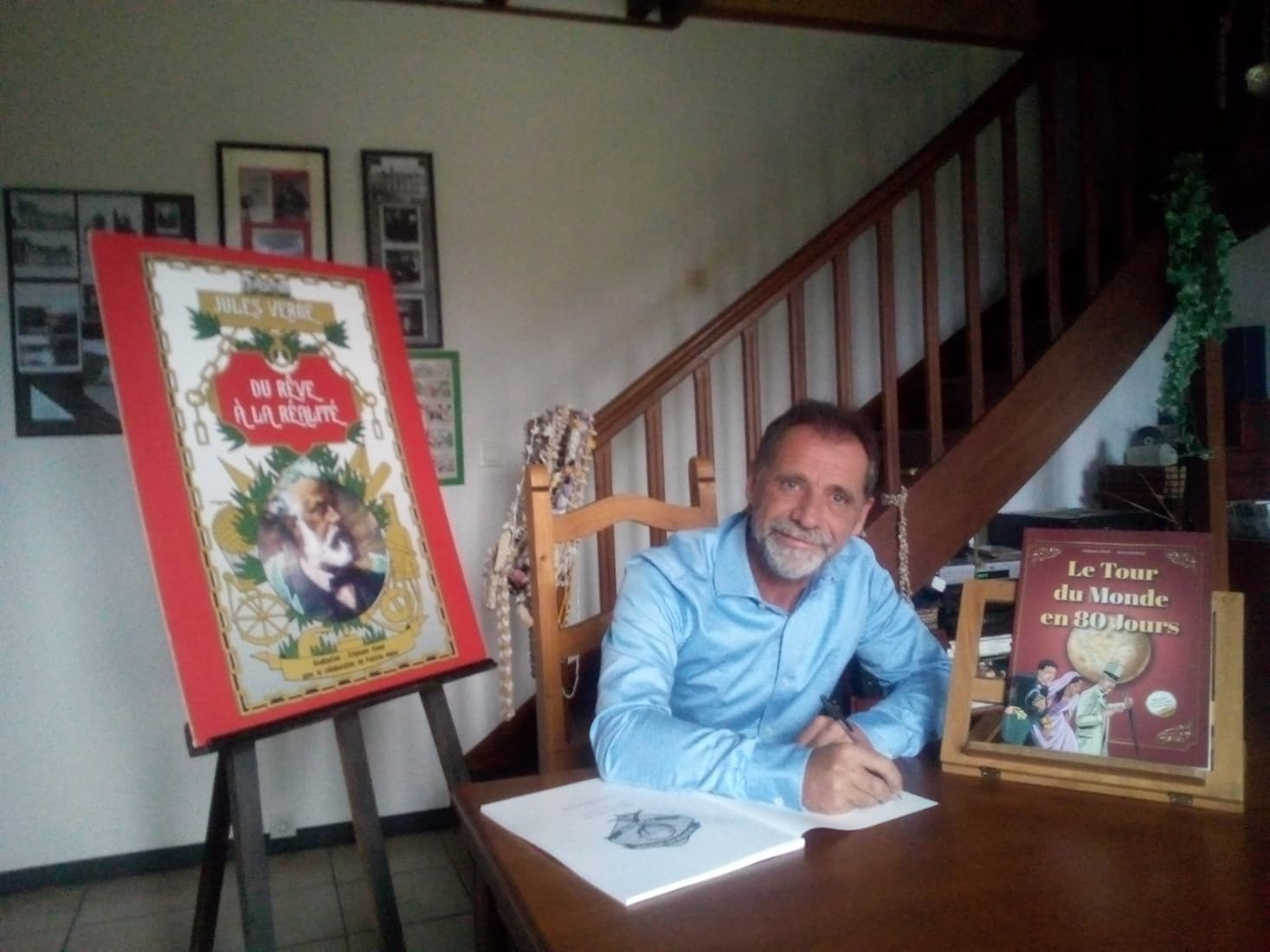 Un homme assis à une table présentant son livre et l'affiche de son exposition